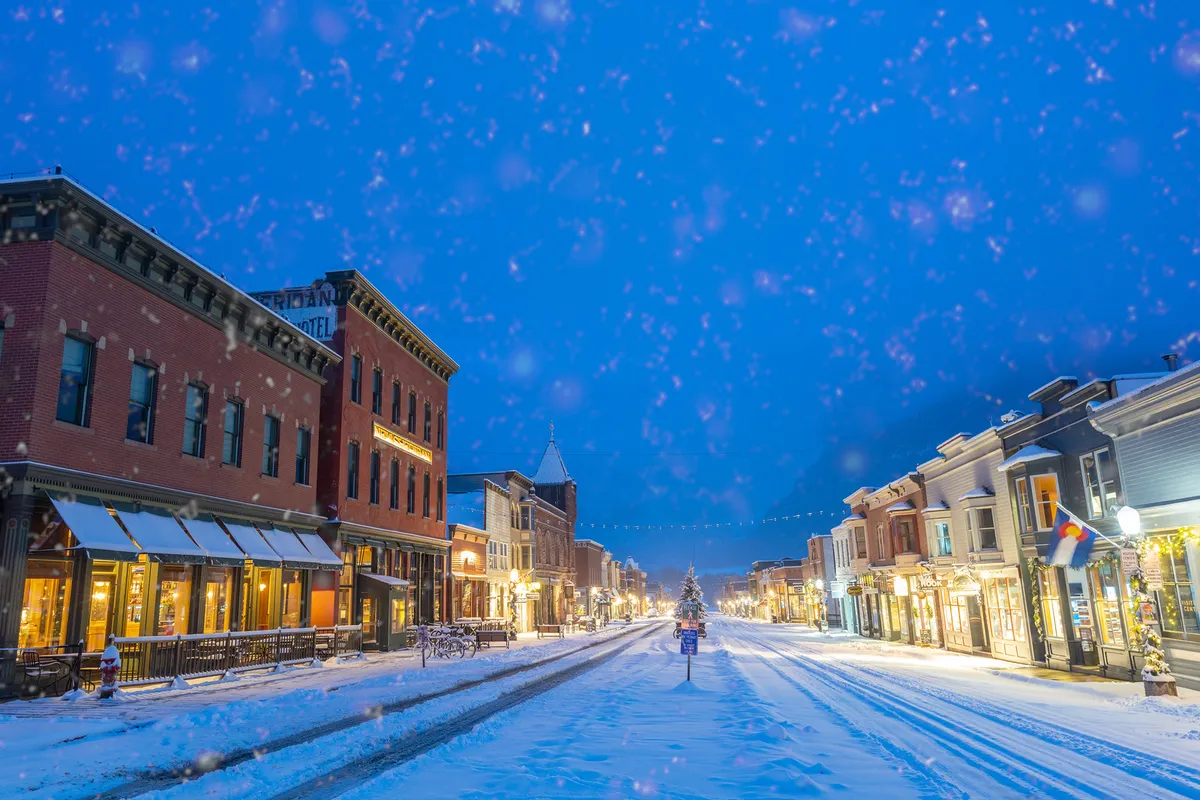 A charming, snow-covered Main Street decorated for the holidays, featuring festive shopfronts and a Christmas tree. The scene is vibrant, inviting visitors to stroll, shop, and enjoy the seasonal ambiance.