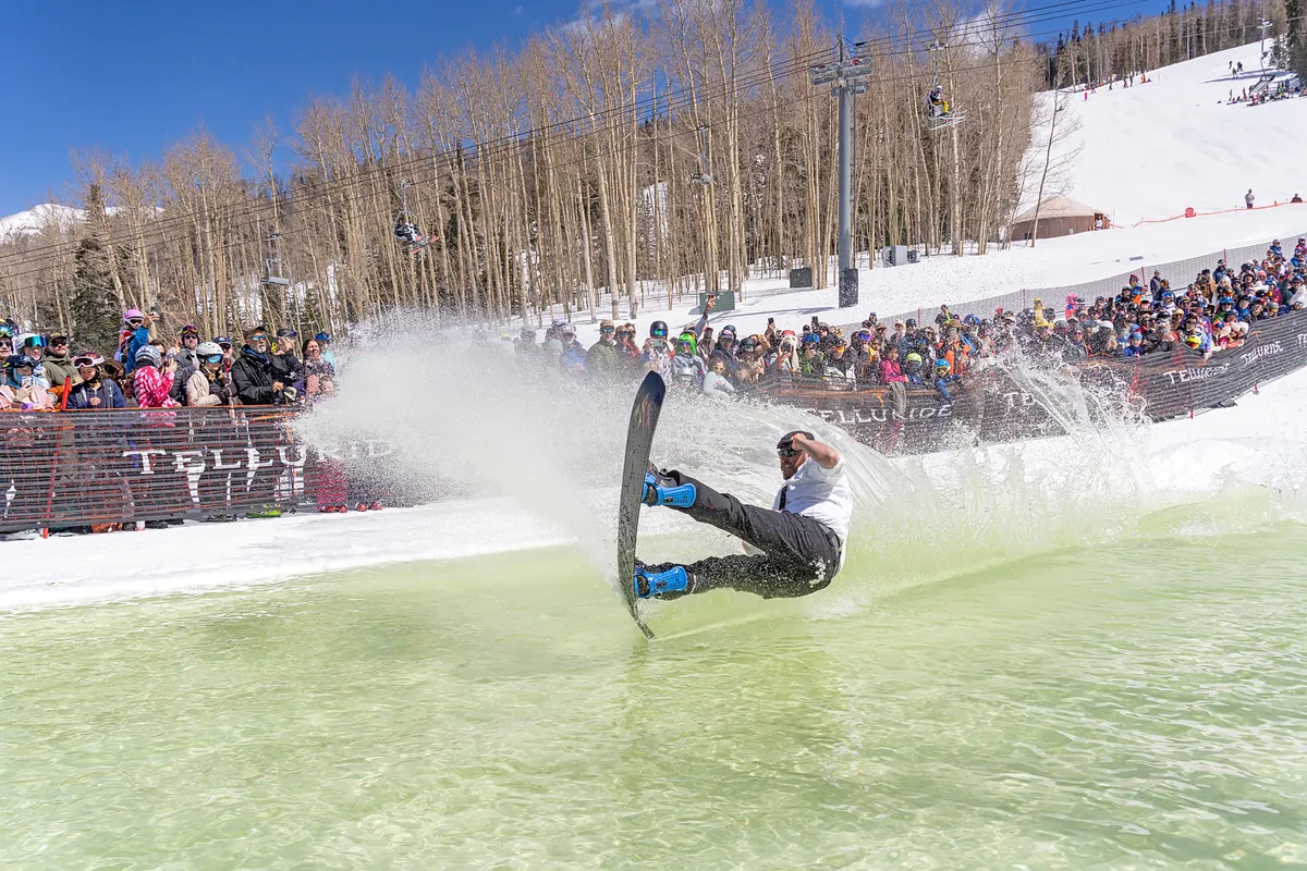 An action-packed exterior shot of a snowboarder performing a trick while spectators cheer, with a dramatic splash of water. The image pulses with excitement and showcases a communal, festival-like atmosphere.