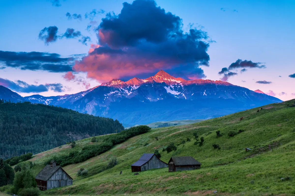 This exterior shot features quaint wooden cabins nestled among lush green hills, with majestic mountain peaks and colorful sunset clouds rising in the background. The scene radiates peace and connection to Telluride's natural beauty, with the cabins suggesting a welcoming home base for visitors.