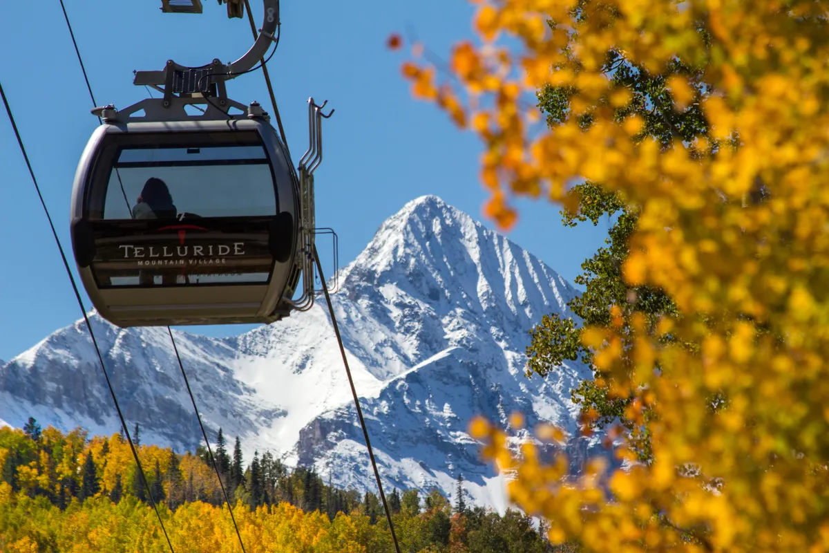 A scenic shot of the iconic Telluride gondola gliding above colorful autumn foliage with snow-capped mountains in the background. The gondola’s presence suggests easy access to both mountain adventures and the heart of downtown. The crisp air and stunning landscape evoke the area’s mild climate and outdoor lifestyle.