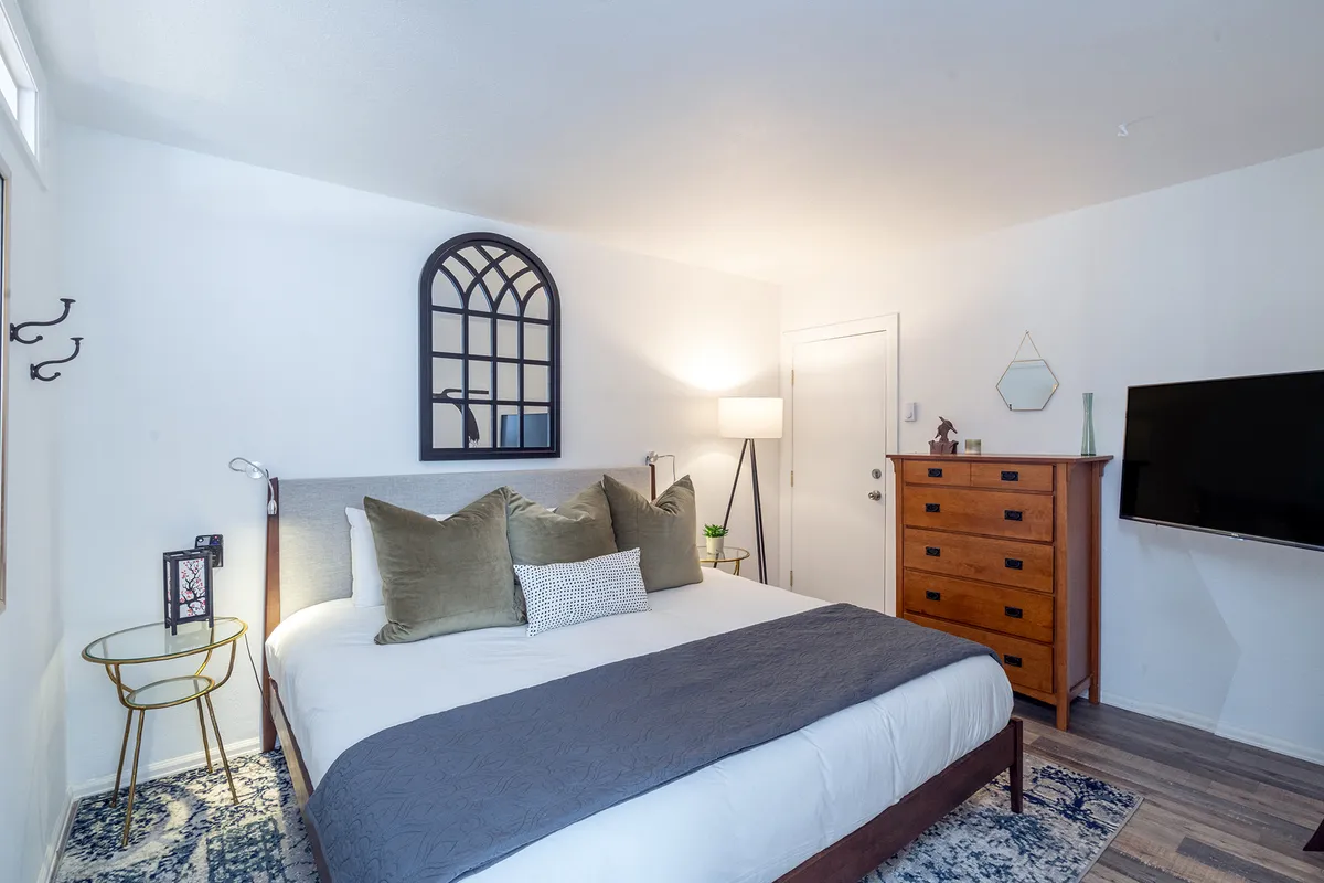 This image features a serene bedroom with a plush bed adorned with decorative pillows, an arched mirror, a wooden dresser, floor lamp, and a flat-screen TV. The soft morning light filters in, creating a soothing, restful atmosphere that invites relaxation and quiet moments.