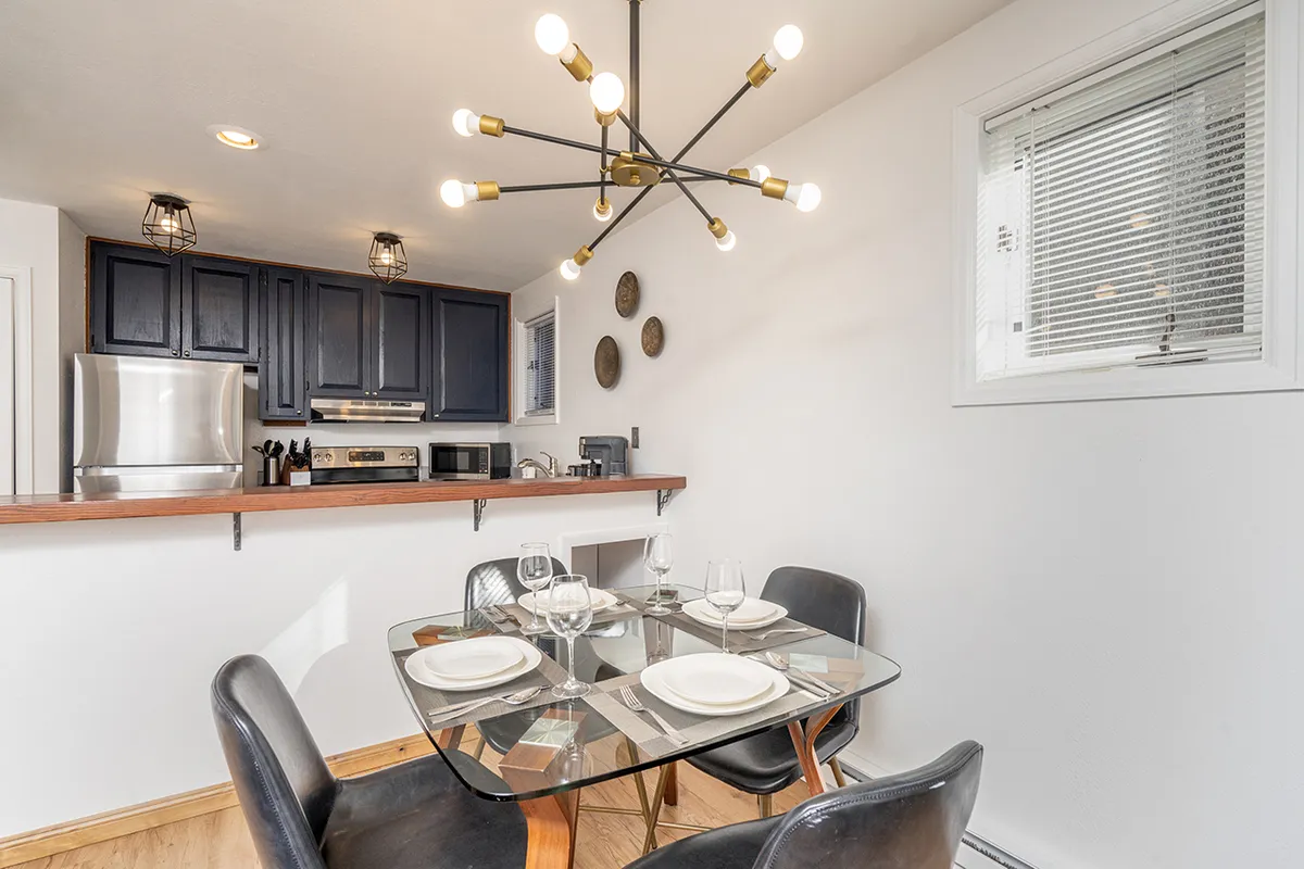 A view of the open-plan dining and kitchen area, featuring a central dining table beneath a modern chandelier, stylish cabinetry, and stainless steel appliances. The kitchen is well-equipped and seamlessly flows into the dining space, suited for group meals and social interaction.