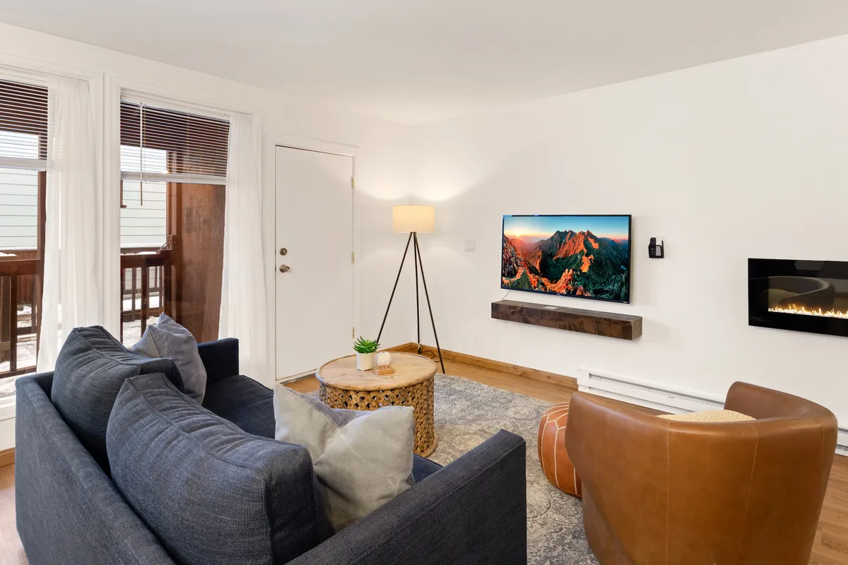 A comfortable, modern living room featuring a cozy sofa, coffee table, TV mounted above an electric fireplace, and a lamp in the corner, all awash in natural light. Clean lines, contemporary finishes, and a welcoming atmosphere make it perfect for relaxing or streaming a movie after a day outdoors.
