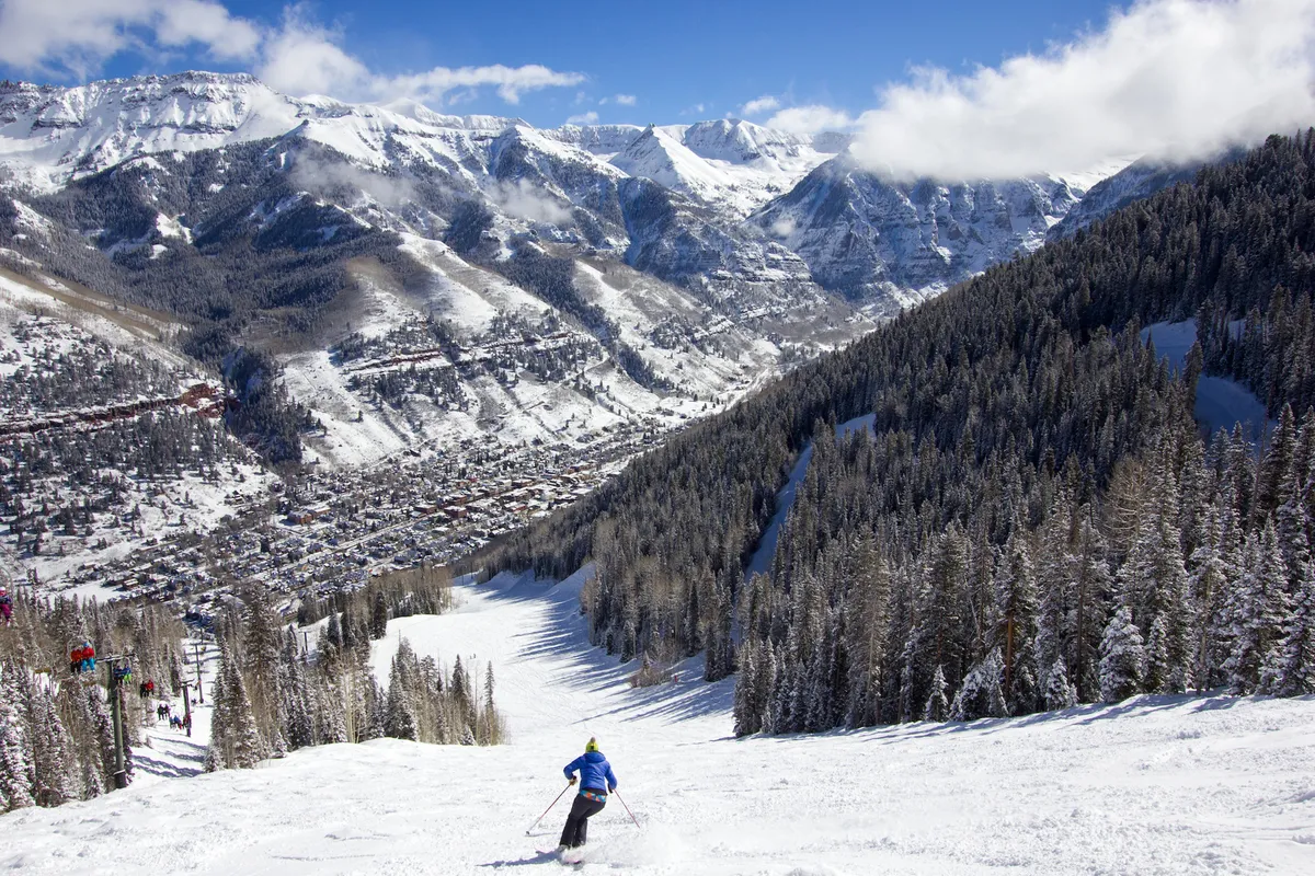 A skier gracefully descending a snowy slope, framed by pine trees and overlooking the town nestled in a valley below. The panoramic backdrop of snow-covered mountains showcases Telluride’s natural beauty and sense of adventure.