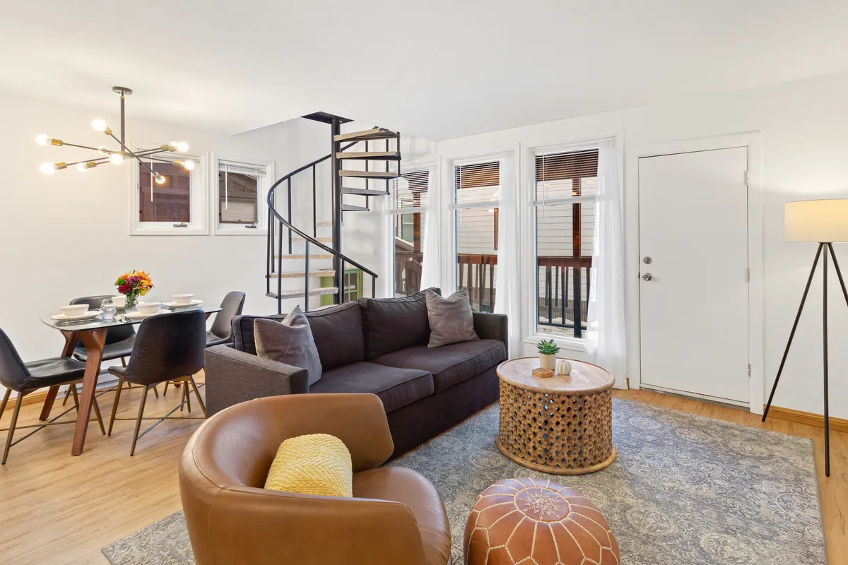 The image showcases a bright, open-plan living and dining space with a stylish spiral staircase as a focal point. A modern dining table invites intimate meals, while a cozy sofa and coffee table are arranged for relaxation or reading by the electric fireplace. The setting exudes warmth, comfort, and the sense of a welcoming mountain retreat.