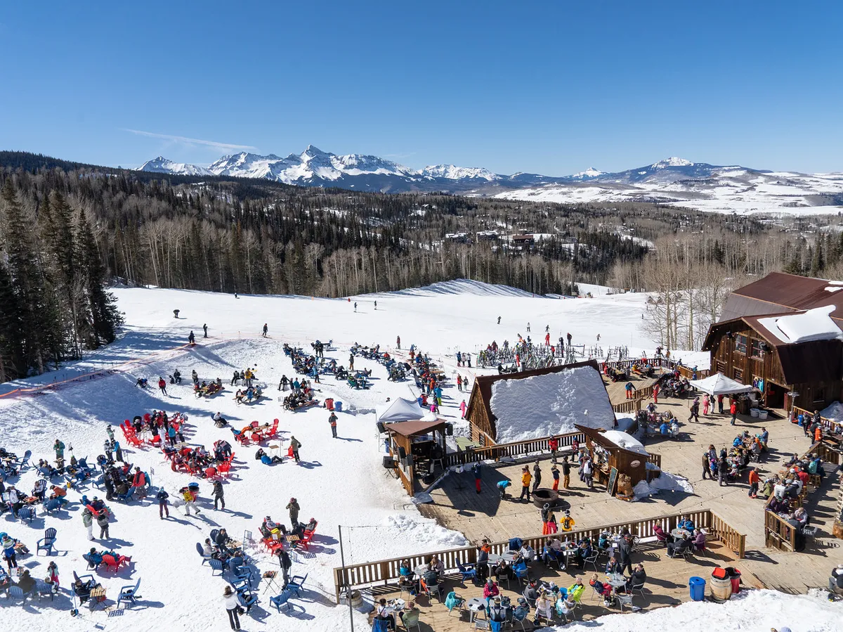 A lively scene at the base of a ski slope, with people socializing on a wooden deck, colorful chairs, and majestic mountains rising in the background. The crowd exudes joy, many with ski gear, and the area feels festive and welcoming.