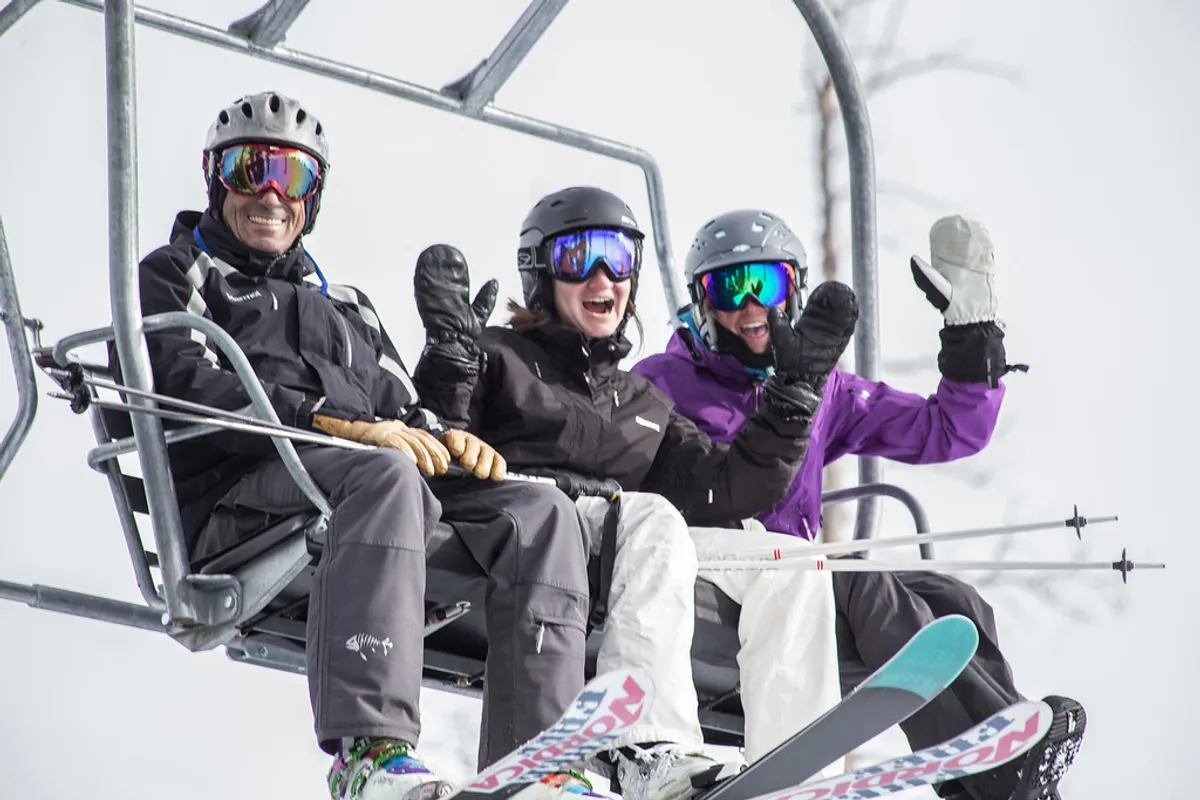 Three skiers ride a chairlift high above a snow-covered mountain landscape. They wear colorful ski gear, and the background showcases dramatic peaks and blue sky, capturing the thrill and camaraderie of a day on the slopes.