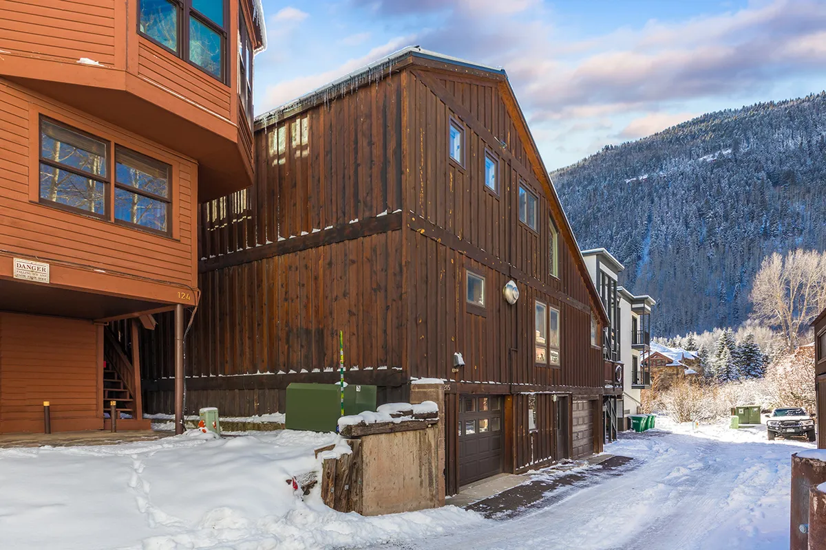 A welcoming wooden building is nestled amidst a snowy landscape, with a dramatic mountain backdrop that hints at the proximity to winter sports and adventure. Fresh snow covers the ground, creating an inviting atmosphere for skiers and snowboarders. The architecture feels cozy and alpine-inspired, complementing the mountain setting.