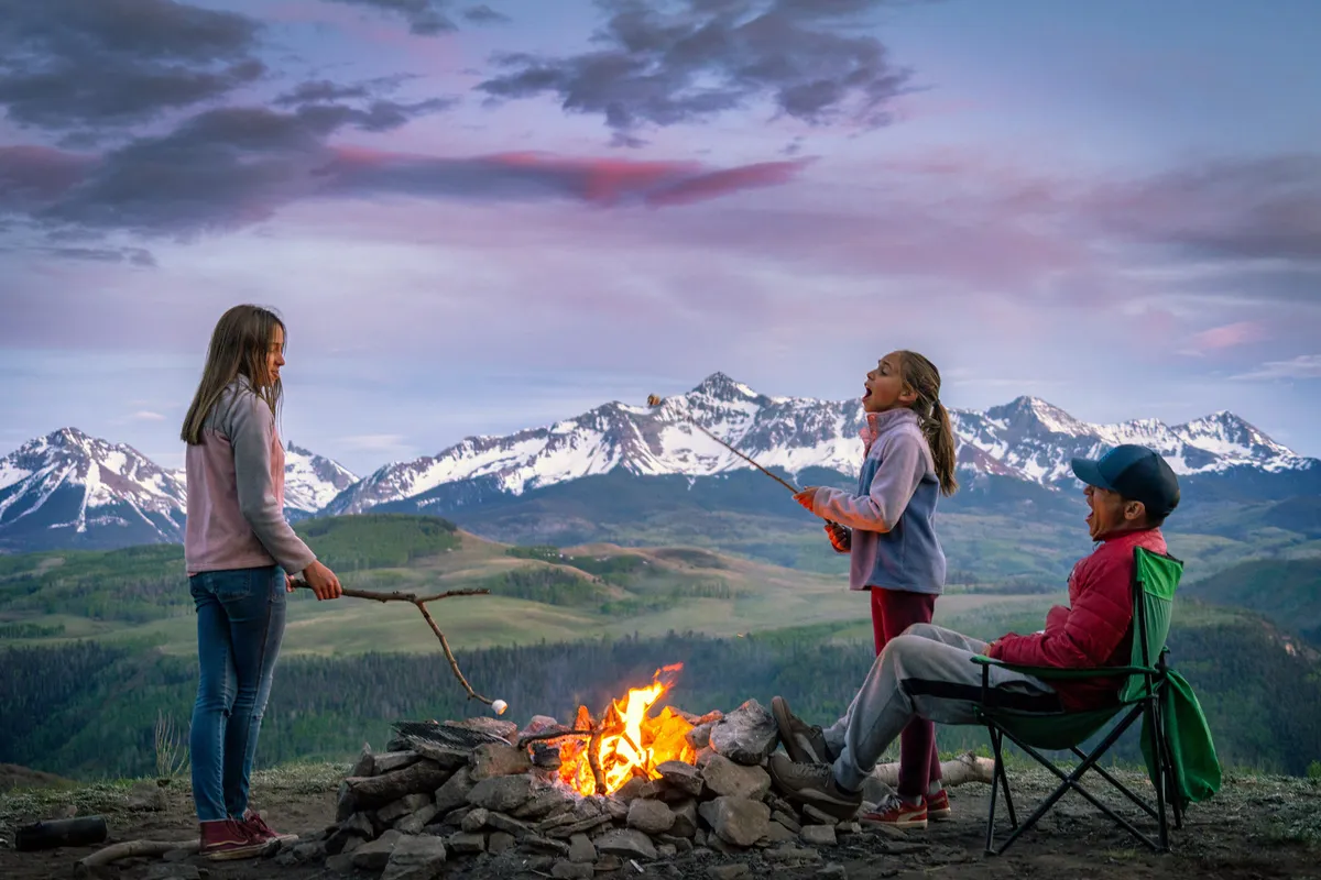 This image captures children roasting marshmallows over a campfire, set against a sweeping mountain landscape at sunset. The joyful scene highlights outdoor family bonding and relaxation, with kids and adults enjoying the natural surroundings together.