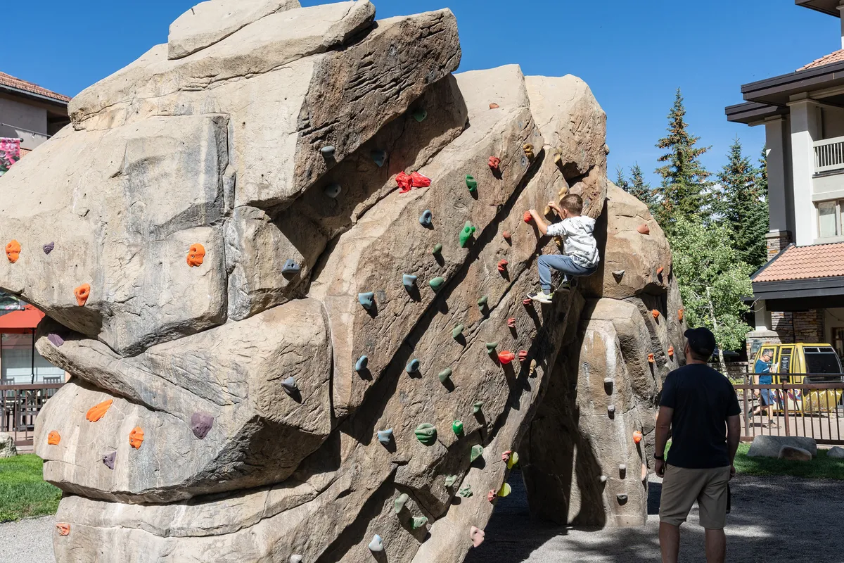 A child climbs a colorful outdoor climbing wall while an adult cheers them on, set in a vibrant, sunny park environment. The image radiates playful energy and family interaction, capturing a special moment of achievement and connection.