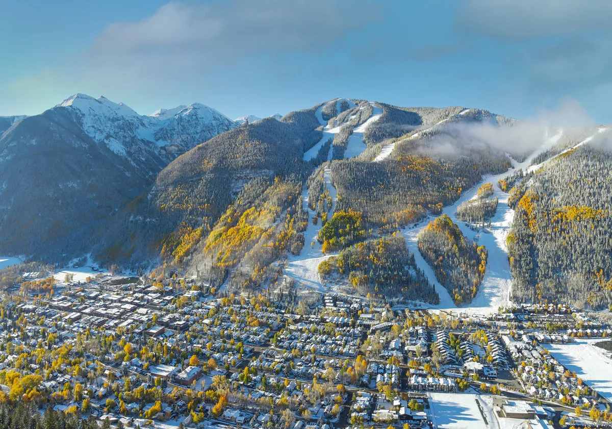 A sweeping aerial or elevated view of Telluride showing snow-capped mountains, colorful autumn trees, ski slopes, and the town’s layout. The image exudes a sense of adventure and tranquility, highlighting the town’s close-knit relationship with its natural surroundings.