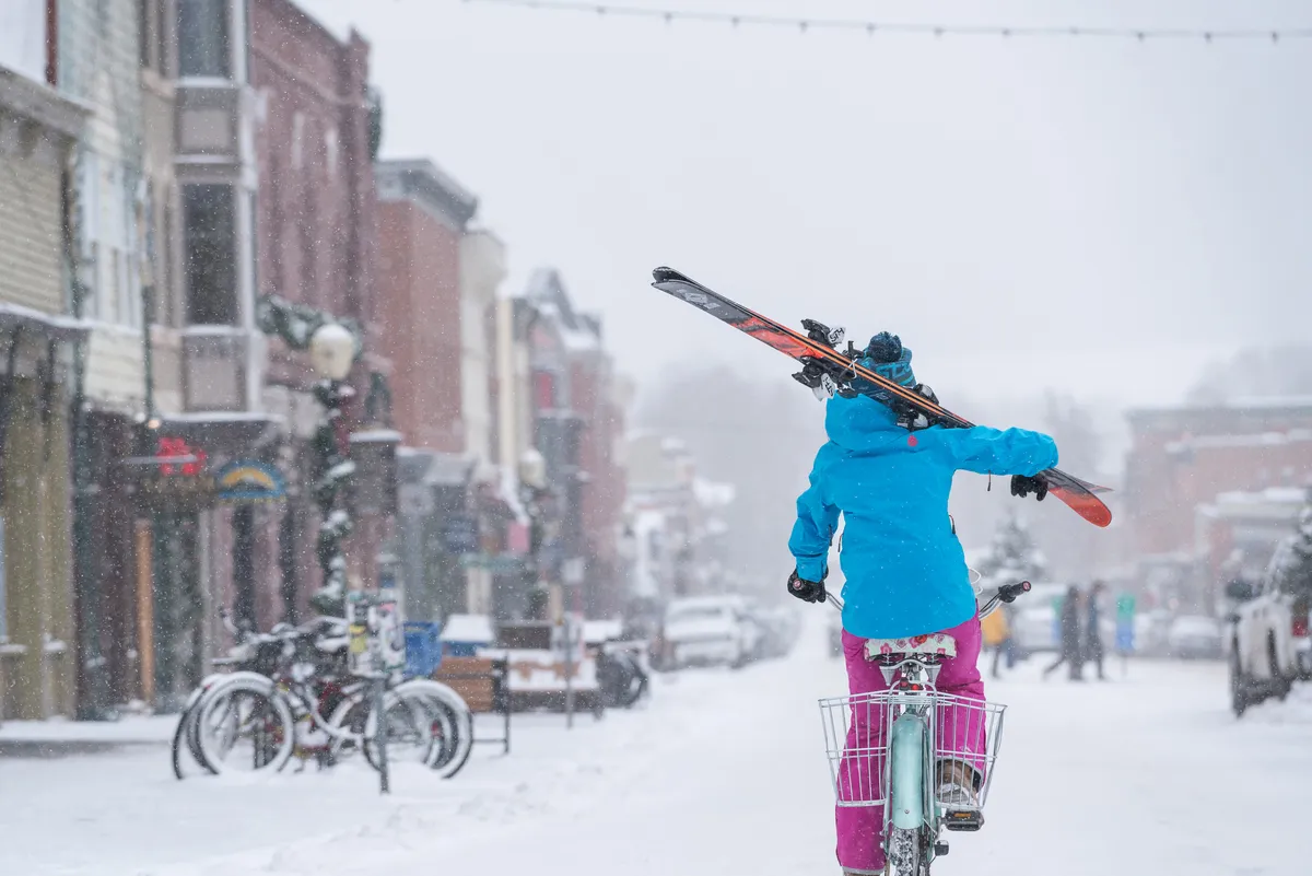 A lively Telluride street scene in winter, featuring a person on a bicycle holding skis overhead. The snow-covered street is lined with historic buildings, and the mood is energetic and joyful, capturing the essence of downtown activity and local culture.