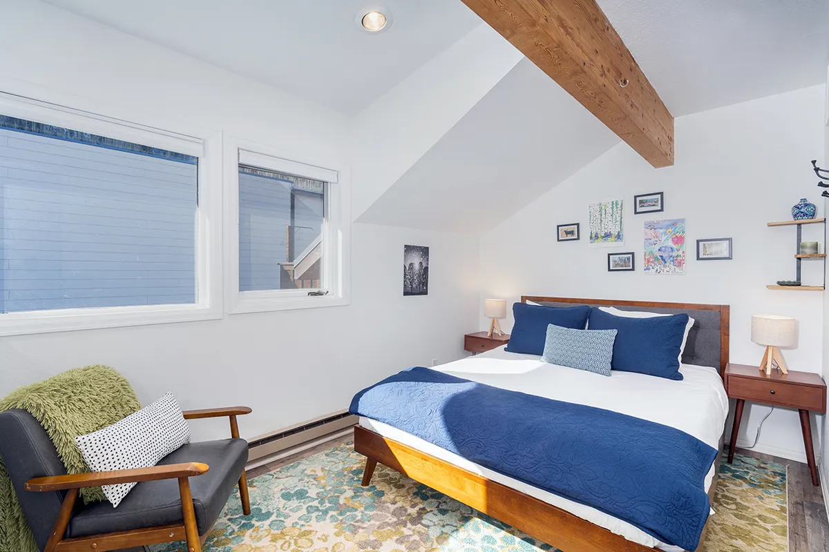 The image shows a loft-like space characterized by a queen-size bed dressed in blue bedding, exposed wooden beams, an armchair, and colorful wall art. Abundant natural light brightens the area, creating a sense of openness and airiness, well-suited for relaxation or reading.
