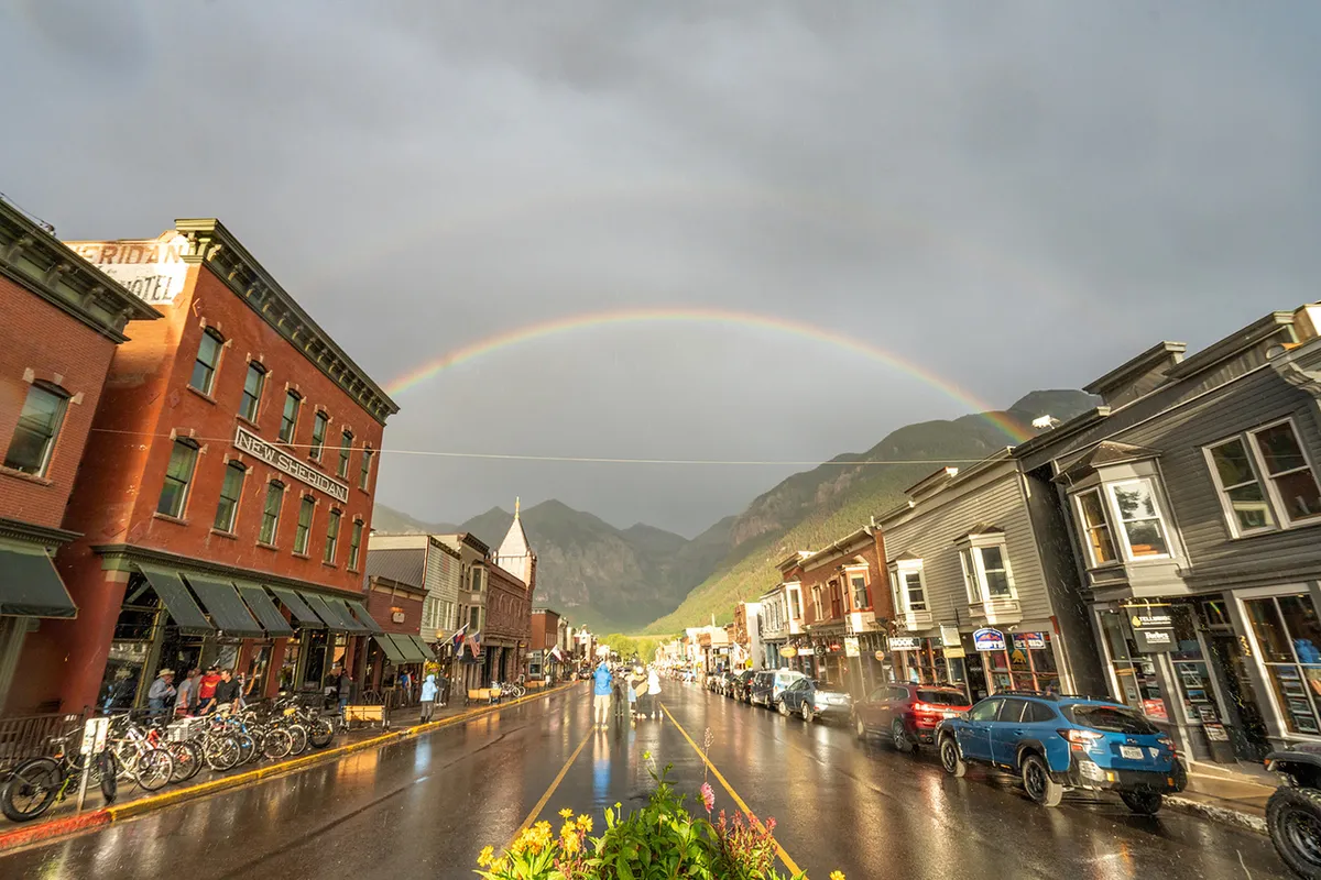 A rainbow arches over a lively main street lined with historic buildings, people strolling, and bicycles parked along the sidewalk. The mood is bright and inviting, with the street scene hinting at festival days and community gatherings.